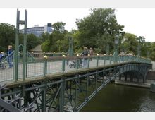 Eine schmiedeeiserne Fußgängerbrücke mit grünem Geländer und goldenen Verzierungselementen überspannt einen Fluss. Im Hintergrund sind Bäume und ein modernes Gebäude erkennbar, während Personen mit Kinderwagen und Fahrrädern die Brücke nutzen.