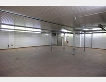 Großer, leerer Raum mit weiß gefliesten Wänden, grauen Metallstützen und einer abgehängten Decke aus Metallpaneelen. Der Boden ist mit rötlich-braunen Fliesen bedeckt und es sind Ablaufgitter sichtbar; an der Wand befinden sich eine Metalltür und ein Feuerlöscher.