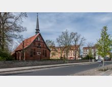 Backsteinkirche mit spitzem Turm und roten Dachziegeln, umgeben von Bäumen und einer niedrigen Mauer; im Hintergrund stehen mehrstöckige Wohnhäuser mit hellen Fassaden. Eine asphaltierte Straße und ein Gehweg verlaufen im Vordergrund.