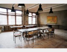 Großer, heller Raum mit industriellem Design, einer Kombination aus rohen Wänden, Holzboden und großen Fenstern mit Jalousien. Zentral stehen mehrere zusammengestellte Tische, umgeben von verschiedenen Stühlen, dazu hängen schwarze Hängeleuchten von der Decke; an der Wand hängt eine Karte.