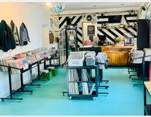 Ein Plattenladen mit türkisfarbenem Boden, gefüllt mit Regalen und Wägen voller Vinyl-Schallplatten. Im Hintergrund befindet sich eine Theke mit Holzfront, dahinter schwarz-weiße Wandstreifen, hängende Lampen und Pflanzen. An einer Wand hängen Jacken, und oben hängt eine Discokugel.