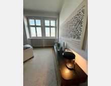 Schmales Schlafzimmer mit großen, mehrteiligen Fenstern und Heizkörper darunter, hellem Teppichboden und einem Bett am linken Rand. Entlang der rechten Wand steht eine lange Kommode mit Lampe und Dekoration, darüber hängt ein großes gerahmtes Poster.