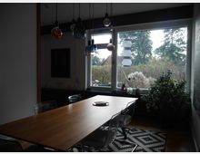 Esszimmer mit einem Holztisch und sechs transparenten Stühlen, beleuchtet von mehreren Pendelleuchten in verschiedenen Farben und Formen. Im Hintergrund ist ein großes Fenster mit Blick auf einen Garten mit Bäumen und Sträuchern, daneben Zimmerpflanzen und Dekorationen auf der Fensterbank.