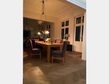 Esszimmer mit hölzernem Esstisch und gepolsterten Stühlen auf einem Fischgrätenparkettboden. Im Hintergrund sind große Fenster, weiße Wände, und eine moderne Hängelampe sichtbar; Kerzen und Dekorationen befinden sich auf dem Tisch.