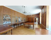 Ein holzgetäfelter Raum mit hellbraunem Parkettboden, schlichten Kronleuchtern und einem langen Tisch aus Holz in der Mitte. Die Wandverkleidung enthält kunstvolle Malereien mit Tier- und Pflanzenmotiven, und im Hintergrund führt eine Tür in einen weiteren Raum.