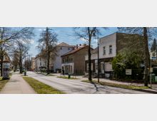 Straßenszene mit einer ruhigen, zweispurigen Straße, flankiert von Gehwegen, Bäumen und Häusern mit einfachen Fassaden. Auf der rechten Seite befindet sich eine Bushaltestelle und links Werbesäulen entlang des Gehwegs.