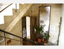 Treppenhaus in einem alten Gebäude mit freiliegenden Ziegelwänden und einem Metallgeländer. Unten im Bild sind eine offene Tür, Topfpflanzen und Poster mit Informationen zu Tanzkursen und Workshops sichtbar.