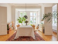 Helle Wohnküche mit einem großen Esstisch, der mit einem weißen Tuch bedeckt ist, umgeben von Holzstühlen. Der Raum hat hohe Decken mit Stuckverzierung, ein großes Fenster, durch das Tageslicht einfällt, Holzboden und dekorative Pflanzen.