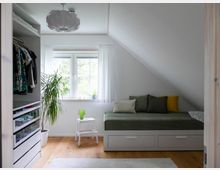 Helles Schlafzimmer mit Dachschräge, einem Tagesbett mit Stauraum und bunten Kissen am rechten Rand. Links ein offener Kleiderschrank und eine Topfpflanze, in der Mitte ein Fenster mit weißem Vorhang, darunter ein kleiner Tisch mit einer grünen Vase.