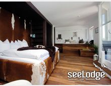 Modern eingerichtetes Hotelzimmer mit einem großen Bett mit Fellbezug, Holzfußboden und einer Sitzecke im Hintergrund. Große Fenster auf der rechten Seite lassen viel Tageslicht herein.