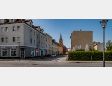 Straßenszene mit einem Eckgebäude, das im Erdgeschoss ein Reisebüro beherbergt, sowie einer Straße, die von weiteren Wohn- und Geschäftsgebäuden gesäumt wird. Im Hintergrund ist eine Kirche mit spitzem Turm sichtbar, während eine Hecke auf der rechten Seite einen Teil des Straßenrandes einnimmt.