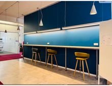 Ein moderner Büroraum mit einer langen, hohen Theke entlang einer blau gestrichenen Wand. Drei Barhocker in Gelb und Schwarz stehen vor der Theke, die durch Hängelampen und LED-Leuchten beleuchtet wird; im Hintergrund ist ein Bürobereich mit Glastrennwänden und einem roten Teppich zu sehen.