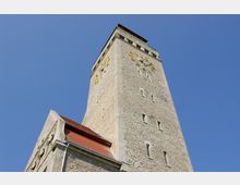 Steinerner Kirchturm mit großer, goldener Uhr an seiner Wand, gesehen aus der Froschperspektive vor klarem, blauem Himmel. Der Turm verfügt über schmale Fenster und ein rotes Ziegeldach an der Seite eines angebauten Gebäudes.