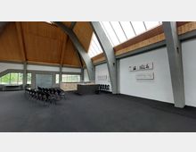 Moderner Saal mit hohen, freitragenden Holzdecken und Betonstützen. Der Raum enthält eine Sitzreihe mit schwarzen Stühlen, einen kleinen Infotresen aus Stein und Glas sowie Informationsplakate an einer weißen Wand; große Fenster lassen Tageslicht herein.