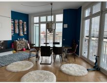 Wohnzimmer mit blau gestrichener Wand, auf der schwebende Bücherregale angebracht sind. Der Raum enthält einen Esstisch mit Stühlen, große Fenster mit Blick auf eine Stadtlandschaft, einen Kamin links sowie helle Holzfußböden mit runden weißen Teppichen.