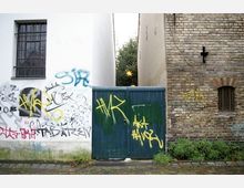 Ein schmaler Hofeingang zwischen zwei Gebäuden, links eine weiß verputzte Wand mit Graffiti, rechts eine Fassade aus Ziegelstein mit einem kleinen Fenster und einem schwarzen Kreuz. Der Eingang wird von einem blauen Tor mit gelben Graffitis verschlossen, davor befinden sich ein gepflasterter Weg und Büsche.