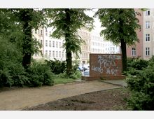 Ein kleiner Stadtpark mit Bäumen und Sträuchern, im Hintergrund eine Reihe von mehrstöckigen Altbauten. Im Vordergrund steht eine rote Backsteinmauer mit Graffiti, daneben ein sandiger Weg.