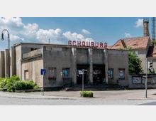 Ehemaliges Kino-Gebäude mit dem Schriftzug „Schauburg“ auf dem Dach, grauer Betonfassade, Plakatresten an den Fenstern und Eingang, umgeben von Gehweg, Grünflächen und Pflastersteinen. Im Hintergrund einige Wohnhäuser mit roten Dächern.