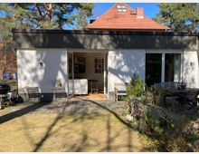 Moderner Bungalow mit Flachdach, weißer Fassade und großen Schiebetüren, die zu einer Holzterrasse führen. Im Vordergrund ein Garten mit Rasen und kleinen Büschen, im Hintergrund ein Haus mit rotem Satteldach und Kiefern.