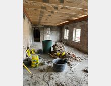 Ein Raum auf einer Baustelle mit freiliegenden Backsteinwänden, unverputztem Boden und einer hölzernen Dachkonstruktion. Auf dem Boden liegen Baumaterialien, wie ein Haufen Ziegelsteine, gelbe Säcke mit Baustoffen, Eimer und Werkzeuge.