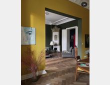 Wohnraum mit einer gelben Wand im Vordergrund, geschmückt mit einem modernen Gemälde und einer Vase mit Trockenblumen. Der Raum öffnet sich zu einem angrenzenden Bereich mit grüner Wand, verziertem Ofen, Stuckaturen an der Decke, Parkettboden in Fischgrätenmuster und einer grauen Polstercouch.