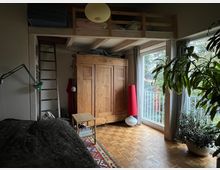 Ein Schlafzimmer mit Holzboden, einem großen hölzernen Kleiderschrank, einer Hochbettkonstruktion mit einer Leiter und großen Fenstern, die auf einen Balkon und grüne Bäume blicken. Eine große Zimmerpflanze steht vor den Fenstern, und eine rote Stehlampe ist in der Ecke zu sehen.