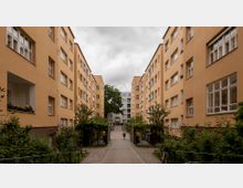 Mehrstöckige Wohngebäude mit gelblichen Fassaden säumen einen gepflasterten Innenhof, der von Bäumen und gepflegten Grünflächen umgeben ist. Im Hintergrund befindet sich ein weiteres modernes Gebäude, während eine Person den Weg entlanggeht.