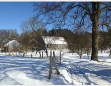 Schneebedeckte Landschaft mit einer kleinen Steinbauernhof-Siedlung im Hintergrund, umgeben von kahlen Bäumen. Im Vordergrund ein verschneiter Pfad, der durch einen Zaun mit Holzbalken verläuft.