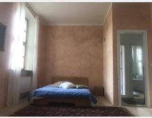 Schlafzimmer mit hellorangefarbenen Wänden, einem niedrigen Holzbett mit blauer Bettdecke und Kissen. Ein großes Fenster mit weißen Vorhängen lässt Tageslicht herein, und eine offene Tür führt zu einem angrenzenden Badezimmer.