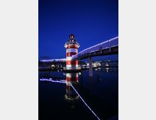 Ein rot-weiß gestreifter Leuchtturm mit einer umlaufenden Aussichtsplattform steht auf einer Wasserfläche und ist über eine beleuchtete Fußgängerbrücke verbunden. Im Hintergrund sind beleuchtete Gebäude und der klare Nachthimmel sichtbar.