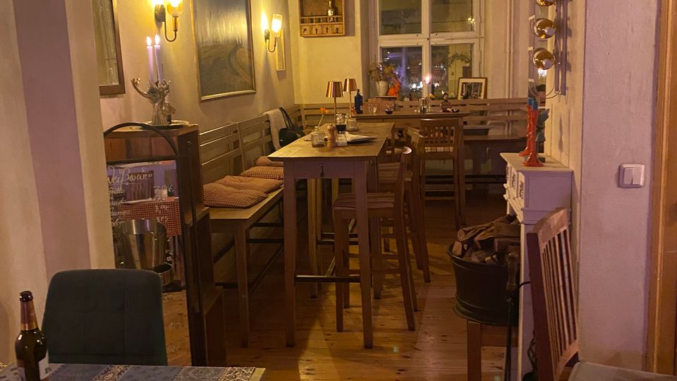 Schmaler, gemütlicher Gastraum mit Holzdielenboden, langen Holzbänken und hohen Tischen, an den Wänden große Bilder und warme Wandleuchten. Am Ende des Raums befindet sich ein Doppelfenster, daneben Regale und verschiedene Dekorationselemente.