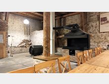 Rustikaler Innenraum mit gemauerten Wänden, freiliegenden Holzbalken und einem großen schwarzen Kaminofen auf einem erhöhten Ziegelpodest. Im Vordergrund stehen Holztische und -stühle, im Hintergrund eine Holzlagerstelle.