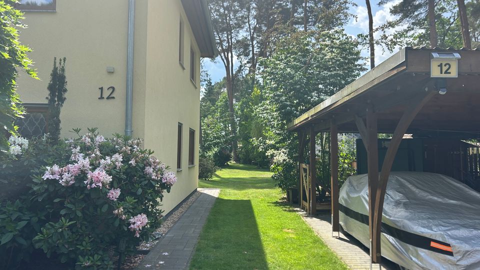 Seitlicher Blick auf ein helles Wohnhaus mit der Hausnummer 12, neben einem gepflasterten Weg und blühenden Sträuchern. Rechts steht ein hölzernes Carport mit überdachtem Stellplatz, dahinter ein grüner Garten mit hohen Bäumen.