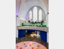 Innenansicht einer umgestalteten Kirche mit gewölbter Decke und hohen gotischen Fenstern. Der Raum verfügt über eine moderne Inneneinrichtung mit bunten Elementen, darunter eine erhöhte Sitzfläche mit Kissen, Pflanzen, eine Bühne sowie farbige Dekosteine auf einem rosa Teppichboden.