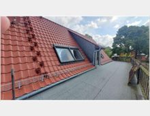 Dachterrasse mit grauem Bodenbelag und einem angrenzenden Schrägdach mit roten Ziegeln und zwei Dachfenstern. Am Dach sind Schneefanggitter angebracht, und im Hintergrund sind Bäume sowie ein Wohngebiet sichtbar.