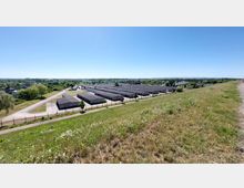 Weitläufiges Gelände mit mehreren flachen, rechteckigen Lagergebäuden, die in Reihen angeordnet sind. Im Vordergrund ein grasbewachsener Hügel, umgeben von vereinzelten Bäumen und Wegen, unter klarem, blauem Himmel.