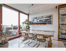 Esszimmer mit großem Holz- und Glastisch, der von Wiener Stühlen umgeben ist. Große Fenster bieten Blick auf eine Terrasse mit Pflanzen, daneben eine Vitrine mit Gläsern und ein modernes Wandbild eines Flugzeugs.