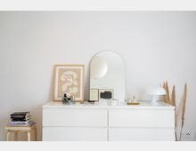 Minimalistisches Schlafzimmer- oder Wohnbereichsdesign mit einer weißen Kommode. Darauf befinden sich ein gerahmtes abstraktes Bild, ein ovaler Spiegel, Bücher, Parfümflakons, eine weiße Tischlampe und dekorative Pampasgräser.