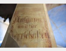 Nahaufnahme eines verwitterten Schriftzugs auf einer sandfarbenen Oberfläche, vermutlich Teil eines alten Gebäudes. Die Schrift ist teilweise unleserlich, zeigt aber Fragmente wie „Eingang... für...“, und im Hintergrund sind unscharf blaue Bodenfliesen erkennbar.