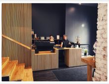 Ein modernes Café mit einer Theke aus Holzlatten, dahinter stehen zwei Personen. Links befindet sich eine Treppe mit Holzstufen, rechts eine weiße Backsteinmauer. Im Hintergrund sind eine Kaffeemaschine, Gläser, und Backwaren sichtbar.