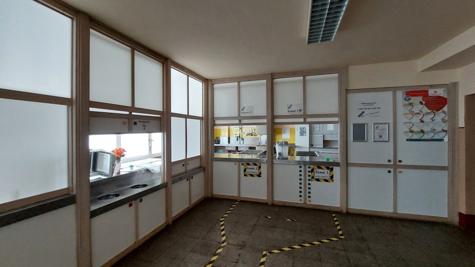 Essensausgabe in einer Kantine mit einer Trennwand aus Milchglas und Holzrahmen. Dahinter sind zwei Essensausgaben mit Schildern ("Essen 1" und "Essen 2"), sowie eine Küche mit gelben Fliesen sichtbar. Auf dem Boden markieren gelbe Klebebänder Laufwege.