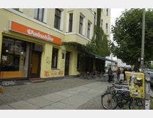 Straßenszene mit einem gelb verputzten Gebäude, das teilweise von Kletterpflanzen überwuchert ist. Im Vordergrund befinden sich ein Café mit Sitzplätzen im Freien, geparkte Fahrräder und ein mit Plakaten beklebter Stromkasten.