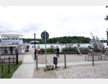 Uferbereich mit einer Bootsverleihstation und einem kleinen Fähranleger an einem See, umgeben von Vegetation und Bäumen. Im Vordergrund stehen Gehwege, Bänke und diverse Skulpturen aus Metall in dynamischen Posen.