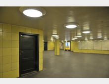 Unterirdischer Korridor mit gelb gekachelten Wänden, schwarzer Tür, leuchtenden Deckenlampen und Werbetafeln an den Wänden. Der Boden ist aus Beton, und an einer Wand sind Karten aufgehängt.