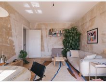 Wohnzimmer mit schlichter, rustikaler Gestaltung: grobe, unverputzte Wände, ein heller Holzboden und moderne Möbel. Im Raum befinden sich ein großes Sofa, ein Couchtisch, ein Regal mit Büchern, eine großblättrige Zimmerpflanze und ein abstraktes Wandbild.