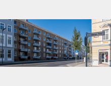 Straßenszene mit mehrstöckigen Wohngebäuden auf der linken Seite, gekennzeichnet durch Balkone und braune Fassaden, sowie einem Straßenschild mit der Bezeichnung „Leipziger Platz“ im Vordergrund. Der Gehweg ist von Bäumen gesäumt, und im Hintergrund sind weitere Gebäude sichtbar.