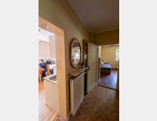 Schmaler Wohnungsflur mit Parkettboden, Wandheizkörper und runden Wandspiegel über einer kleinen Ablage; offene Türen führen links in eine Küche und geradeaus in ein Schlafzimmer mit Bett und Fenster.