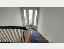 Treppenhaus mit hölzernen Stufen, schwarzem Handlauf und weißen Wänden, das zu einem großen Erker mit mehreren schmalen Fenstern führt, durch die Bäume sichtbar sind.