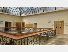 Obergeschoss eines Gebäudes mit einem Glasdach und gelb gestrichenen Wänden. Im Zentrum befindet sich ein Geländer mit dekorativen Metallverzierungen, umgeben von Holztüren, einem Hinweis auf Gebäude B und einer Pinnwand an den Wänden.