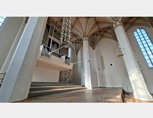 Innenraum einer hohen Kirche mit Rippengewölbe, weißen Pfeilern und großen Spitzbogenfenstern. Links sind eine erhöhte Orgelempore mit sichtbaren Orgelpfeifen und darunter ansteigende Sitzstufen sowie eine freie Bühne im Vordergrund zu sehen.
